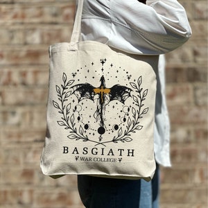 Op de afbeelding: Een beige tote bag met een zwart en gouden ontwerp met een draak, sterren en manen. De tekst "BASGIATH WAR COLLEGE" is op de tas gedrukt.