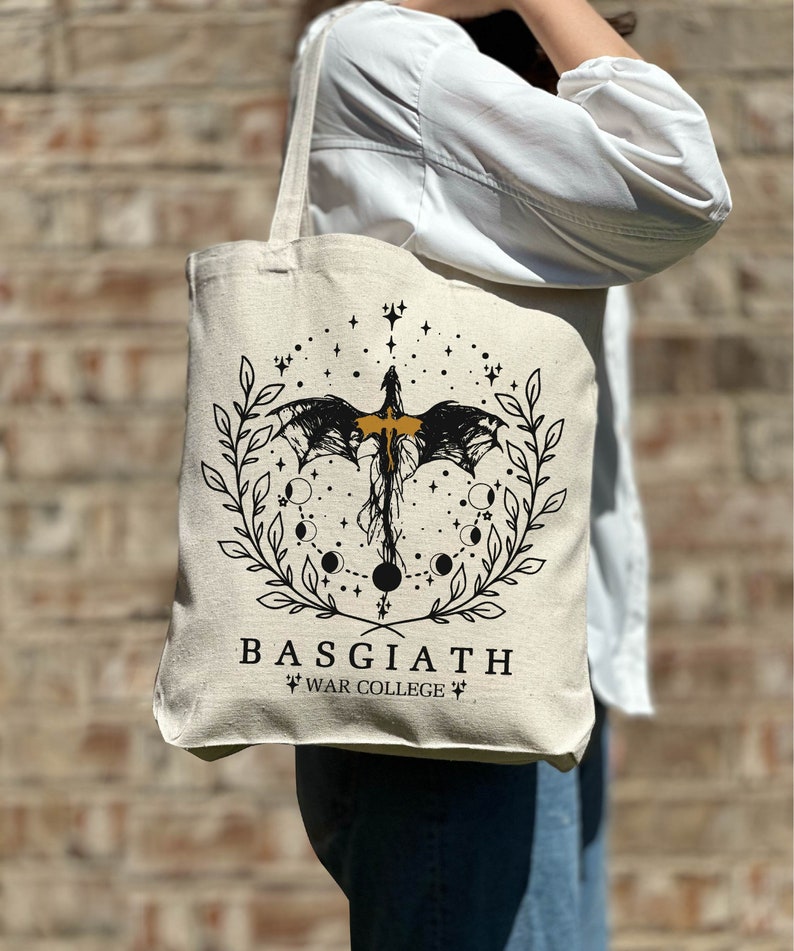 Op de afbeelding: Een beige tote bag met een zwart en gouden ontwerp met een draak, sterren en manen. De tekst "BASGIATH WAR COLLEGE" is op de tas gedrukt.