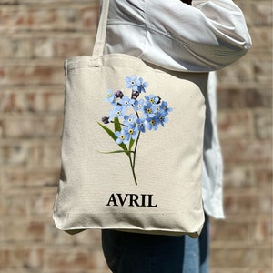 May include: A white canvas tote bag featuring a blue forget-me-not flower design and the word "AVRIL" printed in black.