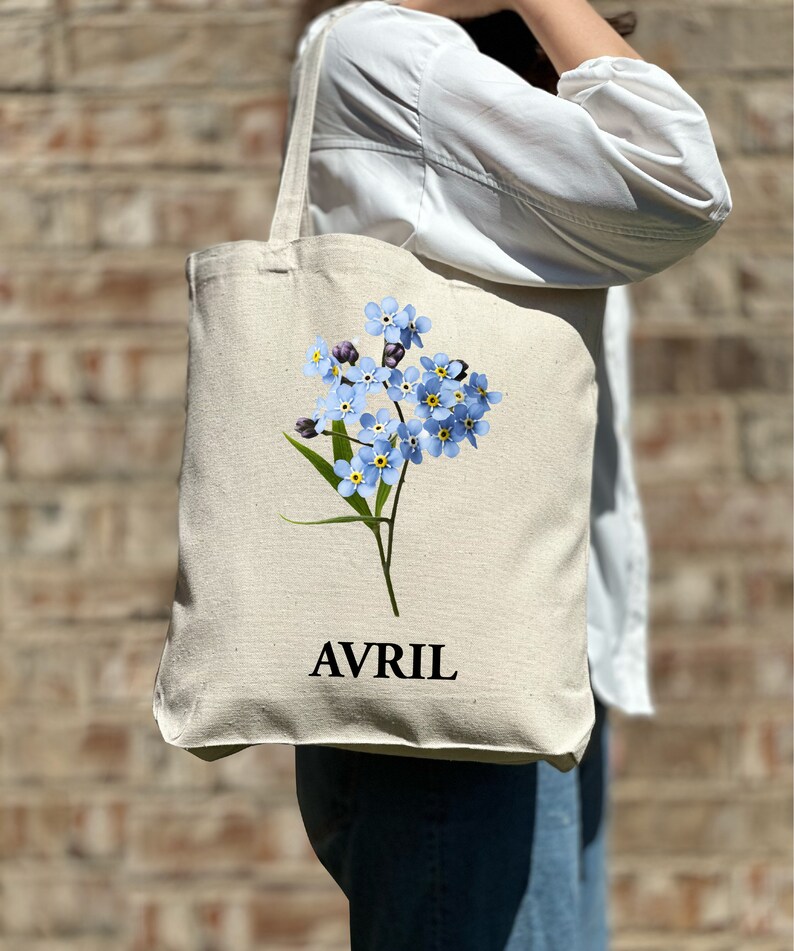 May include: A white canvas tote bag featuring a blue forget-me-not flower design and the word "AVRIL" printed in black.