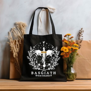 Op de afbeelding: Zwarte tote bag met een wit ontwerp van een draak met uitgespreide vleugels, omringd door een krans van bladeren en manen. De tekst "BASGIATH" en "WAR COLLEGE" is in wit onder het ontwerp gedrukt.