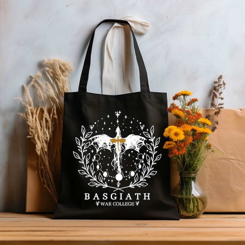 Op de afbeelding: Zwarte tote bag met een wit ontwerp van een draak met uitgespreide vleugels, omringd door een krans van bladeren en manen. De tekst "BASGIATH" en "WAR COLLEGE" is in wit onder het ontwerp gedrukt.