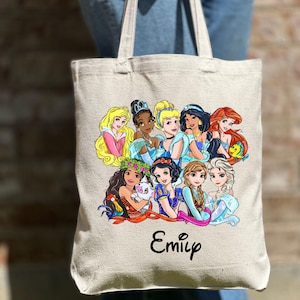 Pu&ograve; includere: Una borsa tote in tela bianca con un'illustrazione colorata di principesse Disney. Le principesse sorridono tutte e si tengono per mano. La borsa ha il nome "Emily" stampato sul davanti.