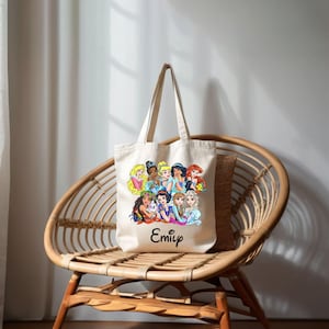 Pu&ograve; includere: Una borsa tote in tela bianca con un'illustrazione colorata di principesse Disney. La borsa ha un manico nero e il nome "Emily" stampato sul davanti.