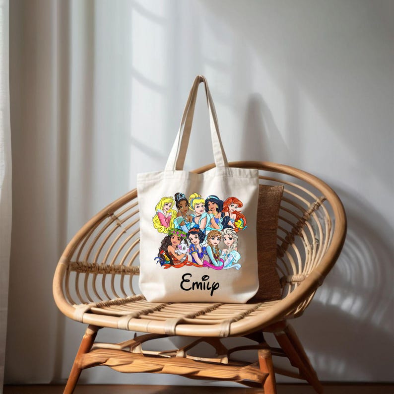 Pu&ograve; includere: Una borsa tote in tela bianca con un'illustrazione colorata di principesse Disney. La borsa ha un manico nero e il nome "Emily" stampato sul davanti.