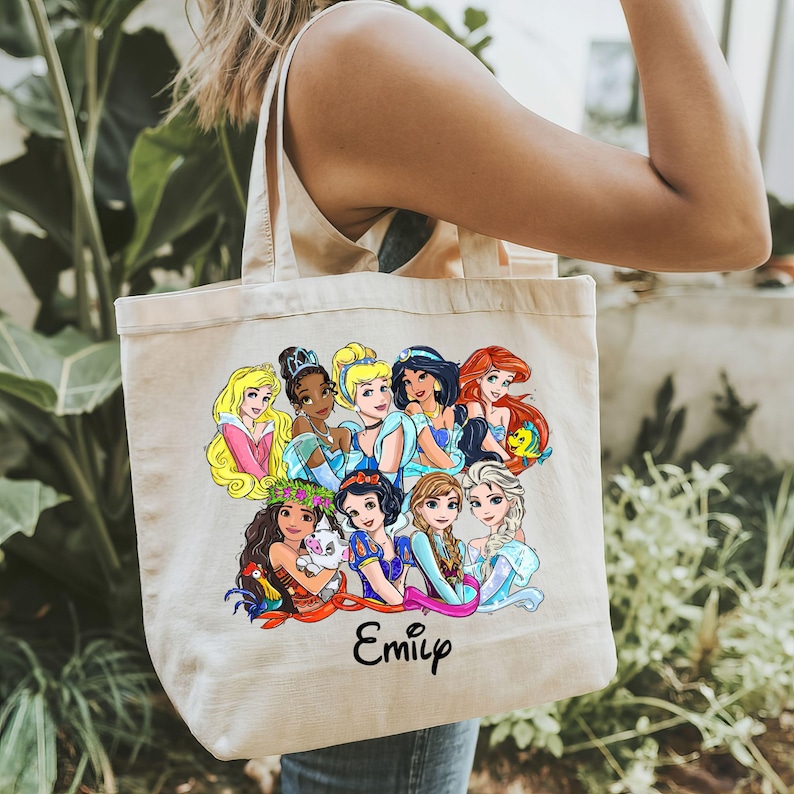Pu&ograve; includere: Una borsa tote beige con un'illustrazione colorata di principesse Disney. Le principesse sono disposte in cerchio con i loro nomi scritti sotto. La borsa ha un nome personalizzato "Emily" stampato sul fondo.