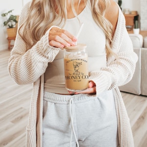 May include: A woman holding a glass jar with a brown label that reads "Hundred Acre Woods" and "Honey Co. Est. 1948".