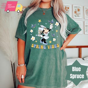 Pode incluir: Uma camiseta verde com uma imagem de desenho animado do Mickey Mouse segurando uma flor e cercada por borboletas e estrelas. O texto "SPRING VIBES" &eacute; impresso abaixo da imagem.