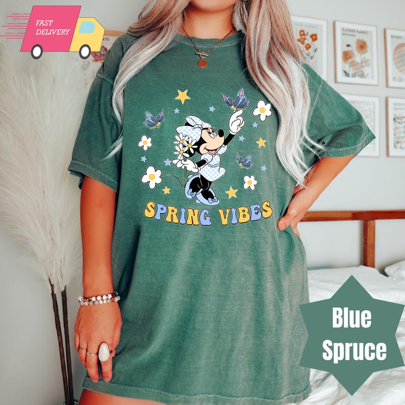 Pode incluir: Uma camiseta verde com uma imagem de desenho animado do Mickey Mouse segurando uma flor e cercada por borboletas e estrelas. O texto "SPRING VIBES" &eacute; impresso abaixo da imagem.
