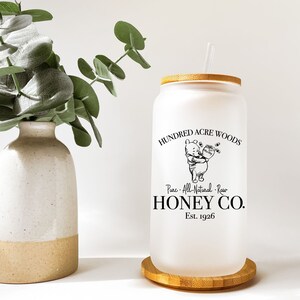 May include: A frosted glass tumbler with a bamboo lid and straw. The tumbler features a Winnie the Pooh illustration and the text "HUNDRED ACRE WOODS Pure. All-Natural. Raw HONEY CO. Est. 1926".
