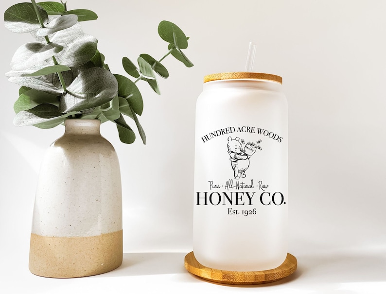May include: A frosted glass tumbler with a bamboo lid and straw. The tumbler features a Winnie the Pooh illustration and the text "HUNDRED ACRE WOODS Pure. All-Natural. Raw HONEY CO. Est. 1926".