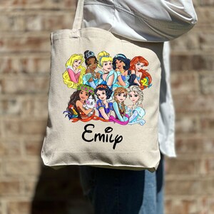 Pu&ograve; includere: Una borsa tote in tela bianca con un'illustrazione colorata di principesse Disney, tra cui Cenerentola, Ariel, Belle e Moana. La borsa ha il nome "Emily" stampato in nero sotto l'immagine.