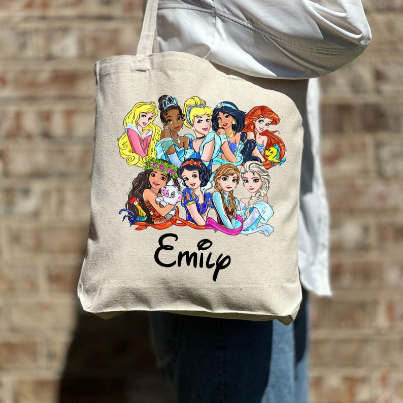 Pu&ograve; includere: Una borsa tote in tela bianca con un'illustrazione colorata di principesse Disney, tra cui Cenerentola, Ariel, Belle e Moana. La borsa ha il nome "Emily" stampato in nero sotto l'immagine.