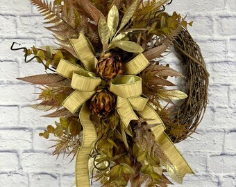 May include: A decorative fall wreath featuring a grapevine base adorned with faux leaves in shades of gold, brown, and green. A large gold ribbon bow and decorative accents complete the design. Perfect for seasonal home decor.