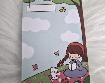 May include: A notepad with a whimsical illustration of a girl reading under a tree. The scene includes a cat, butterflies, and a picnic blanket. The background is a light blue sky with clouds. A blank sign hangs from the tree.