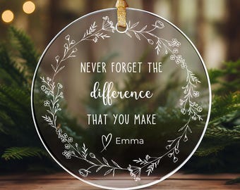 Op de afbeelding: Een ronde, transparante ornament met een wit bloemenontwerp en de tekst "Vergeet nooit het verschil dat je maakt" met een hart en de naam "Emma" eronder.