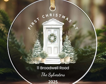 May include: A clear, round Christmas ornament with a gold ribbon. The ornament features a white door with a wreath, flanked by Christmas trees and potted plants. Text reads "First Christmas at 11 Broadwell Road, The Sylvesters, 2025."