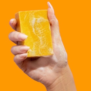 May include: A rectangular, yellow bar of soap held in a hand with long, manicured nails. The soap has a textured surface and is surrounded by white foam, set against a bright orange background.