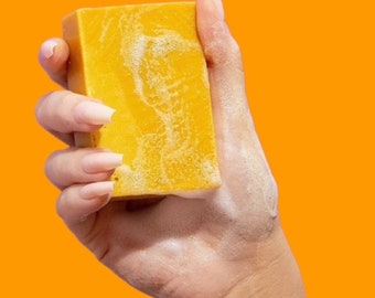 Op de afbeelding: Een rechthoekig, geel stuk zeep in een hand met lange, verzorgde nagels. De zeep heeft een textuur en is omgeven door wit schuim, tegen een heldere oranje achtergrond.