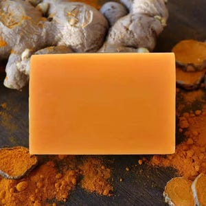 May include: A rectangular bar of solid orange soap positioned centrally. The soap has a smooth, matte surface and uniform color. Surrounding the soap are pieces of raw ginger root and scattered piles of bright orange turmeric powder.