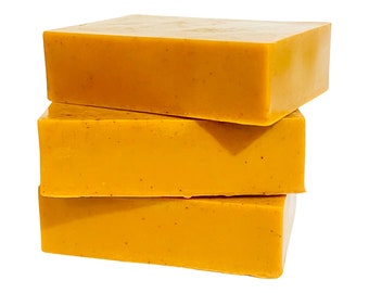 May include: Three rectangular bars of handmade soap stacked. The soap is a vibrant yellow-orange colour with small, dark flecks throughout. The bars have a smooth, slightly glossy surface, suggesting a natural product.
