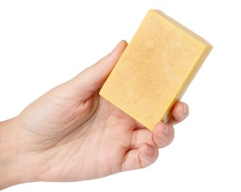 Op de afbeelding: Een hand houdt een rechthoekig stuk gele zeep vast tegen een witte achtergrond. De zeep heeft een licht getextureerd oppervlak en een warme, gouden tint. De zeep lijkt handgemaakt en natuurlijk te zijn.