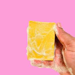 Könnte beinhalten: Ein gelbes Seifenstück mit weißem Schaum, gehalten in einer Hand vor rosa Hintergrund. Die Seife ist mit Blasen bedeckt, was darauf hindeutet, dass sie frisch aufgeschäumt wurde. Die Seife hat eine strukturierte Oberfläche.