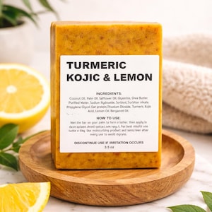 May include: A turmeric and kojic acid soap bar with a white label, on a wooden plate. The label reads "TURMERIC KOJIC & LEMON". A sliced lemon and green leaves are in the foreground. The soap weighs 99 grams.