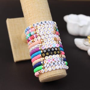 May include: A stack of colorful beaded bracelets with different names spelled out on them, including "Charlie", "River", "Noah", "Dior", "Kate", "Grace", "Jordan", and "Amelia".