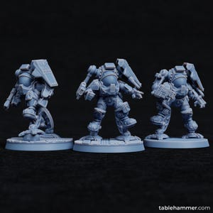 May include: Three light blue miniature figurines of futuristic armored figures. Each figure is posed differently, with detailed armor, helmets, and weapons. The figures are standing on circular bases, and the background is dark.