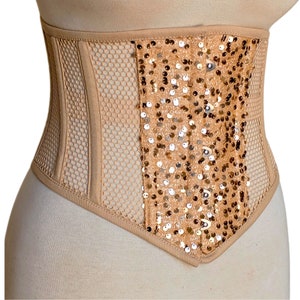 Könnte beinhalten: Ein beiges Korsett mit Mesh- und Pailletten-Design. Das Korsett hat ein zentrales Feld aus schimmernden Pailletten, das von Abschnitten eines Wabennetzes umrahmt wird. Die Kanten sind mit passendem beigem Stoff besetzt, wodurch eine strukturierte Silhouette entsteht.