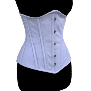 Könnte beinhalten: Ein weißer Korsett mit vertikalen Bahnen und bronzefarbenen Verschlüssen. Das Kleidungsstück wird auf einer schwarzen Schaufensterpuppe präsentiert, um seine Form und sein Design hervorzuheben. Das Korsett hat eine strukturierte Silhouette.