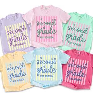 May include: Six short-sleeved t-shirts in lavender, light pink, mint green, pale yellow, light blue, and hot pink. Each shirt features a central rectangular graphic with vertical stripes, small star icons, and the text second grade MISS JOHNSON.