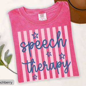 May include: A pink short-sleeved t-shirt with a crew neckline and a Comfort Colors label. The front features a graphic with vertical light pink stripes, dark blue cursive text reading speech therapy, and several dark blue star outlines.