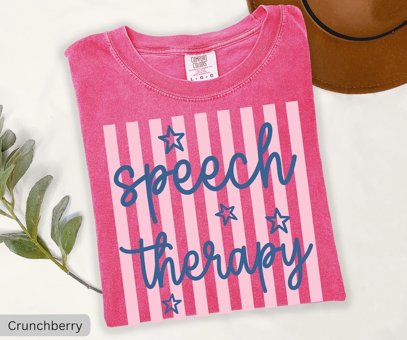 May include: A pink short-sleeved t-shirt with a crew neckline and a Comfort Colors label. The front features a graphic with vertical light pink stripes, dark blue cursive text reading speech therapy, and several dark blue star outlines.