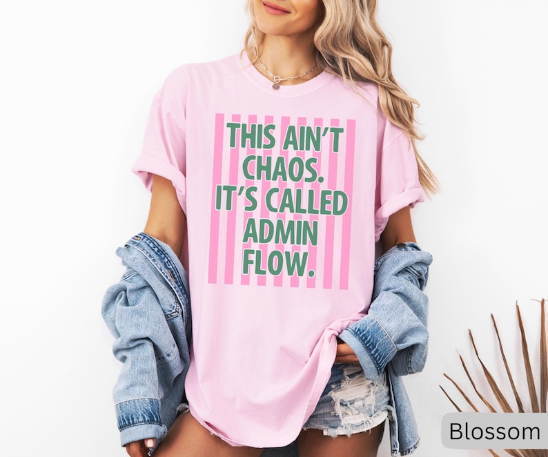 May include: A light pink t-shirt with the text "THIS AIN'T CHAOS. IT'S CALLED ADMIN FLOW." in green and pink. The shirt has a relaxed fit and is paired with a denim jacket and shorts. The shirt is a casual wear item.
