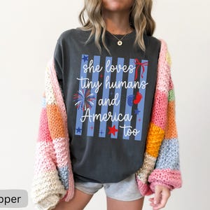 May include: A charcoal gray t-shirt featuring a graphic with vertical light blue stripes and the white text "she loves tiny humans and America too". The design includes red, white, and blue accents such as a firework, a bow, stars, and baby footprints.
