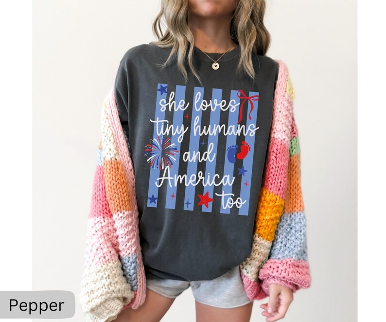 May include: A charcoal gray t-shirt featuring a graphic with vertical light blue stripes and the white text "she loves tiny humans and America too". The design includes red, white, and blue accents such as a firework, a bow, stars, and baby footprints.
