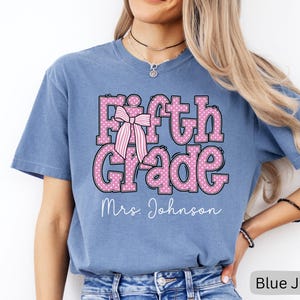 K&ouml;nnte beinhalten: Ein kurz&auml;rmeliges T-Shirt in Jeansblau mit dem Schriftzug Fifth Grade in Pink mit wei&szlig;en Punkten und einer Grafik einer rosa-wei&szlig; gestreiften Schleife. Unter dem Text ist der Name Mrs. Johnson in wei&szlig;er Schreibschrift aufgedruckt.