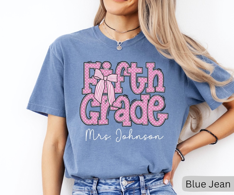 K&ouml;nnte beinhalten: Ein kurz&auml;rmeliges T-Shirt in Jeansblau mit dem Schriftzug Fifth Grade in Pink mit wei&szlig;en Punkten und einer Grafik einer rosa-wei&szlig; gestreiften Schleife. Unter dem Text ist der Name Mrs. Johnson in wei&szlig;er Schreibschrift aufgedruckt.