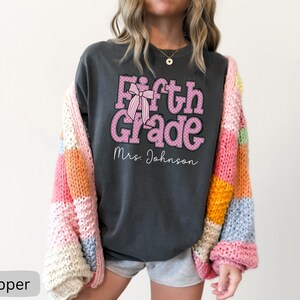 K&ouml;nnte beinhalten: Ein anthrazitfarbenes Kurzarm-T-Shirt mit dem rosa gepunkteten Text Fifth Grade, einer rosa gestreiften Schleifengrafik und dem Namen Mrs. Johnson in wei&szlig;er Schreibschrift, getragen unter einer grob gestrickten Strickjacke mit bunten Patches.