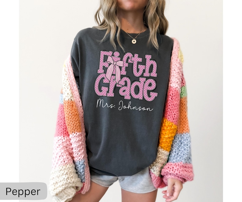 K&ouml;nnte beinhalten: Ein anthrazitfarbenes Kurzarm-T-Shirt mit dem rosa gepunkteten Text Fifth Grade, einer rosa gestreiften Schleifengrafik und dem Namen Mrs. Johnson in wei&szlig;er Schreibschrift, getragen unter einer grob gestrickten Strickjacke mit bunten Patches.