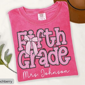 K&ouml;nnte beinhalten: Ein leuchtend pinkes T-Shirt mit Rundhalsausschnitt und dem Text Fifth Grade in gro&szlig;en, wei&szlig; gepunkteten Buchstaben mit schwarzer Umrandung. Eine gestreifte rosa-wei&szlig;e Schleife liegt auf dem Buchstaben F. Der Name Mrs. Johnson steht darunter.