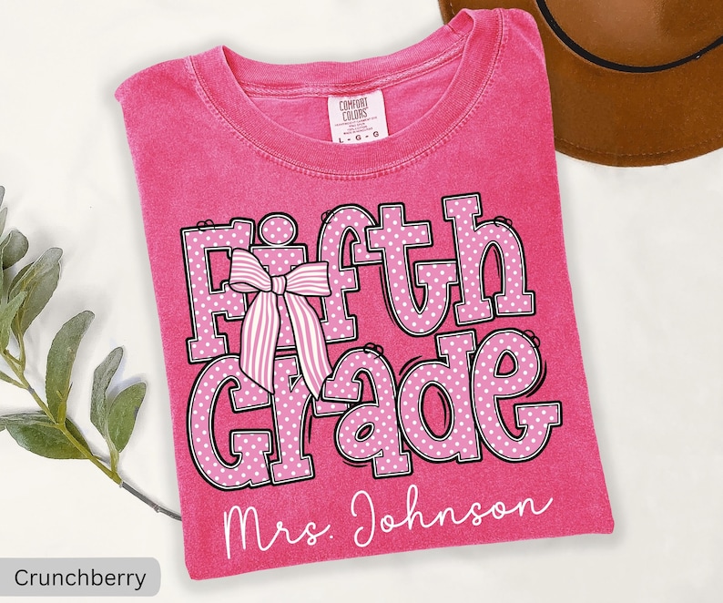 K&ouml;nnte beinhalten: Ein leuchtend pinkes T-Shirt mit Rundhalsausschnitt und dem Text Fifth Grade in gro&szlig;en, wei&szlig; gepunkteten Buchstaben mit schwarzer Umrandung. Eine gestreifte rosa-wei&szlig;e Schleife liegt auf dem Buchstaben F. Der Name Mrs. Johnson steht darunter.