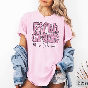 K&ouml;nnte beinhalten: Ein hellrosa T-Shirt mit kurzen &Auml;rmeln, das den Text Fifth Grade in einer rosa-wei&szlig; gepunkteten Schriftart mit einer gestreiften Schleifengrafik zeigt. Unter dem Text ist der Name Mrs. Johnson in einer schwarzen Schreibschrift aufgedruckt.