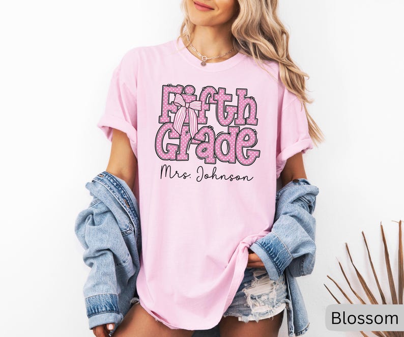 K&ouml;nnte beinhalten: Ein hellrosa T-Shirt mit kurzen &Auml;rmeln, das den Text Fifth Grade in einer rosa-wei&szlig; gepunkteten Schriftart mit einer gestreiften Schleifengrafik zeigt. Unter dem Text ist der Name Mrs. Johnson in einer schwarzen Schreibschrift aufgedruckt.