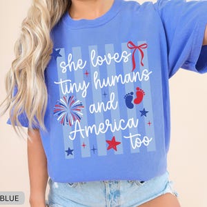 May include: A periwinkle blue short-sleeve t-shirt featuring a graphic print with vertical light blue stripes. The text reads "she loves tiny humans and America too" in white script, accompanied by a red bow, a firework, and baby footprints.