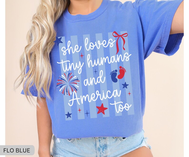 May include: A periwinkle blue short-sleeve t-shirt featuring a graphic print with vertical light blue stripes. The text reads "she loves tiny humans and America too" in white script, accompanied by a red bow, a firework, and baby footprints.