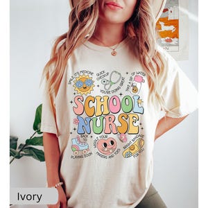 Pode incluir: Camiseta de cor marfim com um gr&aacute;fico colorido que diz "School Nurse" e inclui ilustra&ccedil;&otilde;es de um estetosc&oacute;pio, uma bandagem, uma garrafa de &aacute;gua, um patins e um sol sorridente.