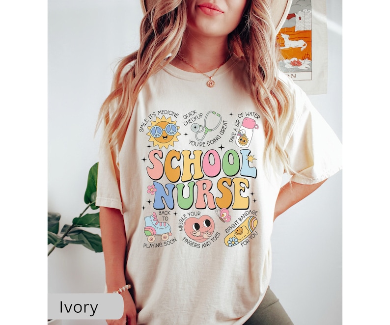 Pode incluir: Camiseta de cor marfim com um gr&aacute;fico colorido que diz "School Nurse" e inclui ilustra&ccedil;&otilde;es de um estetosc&oacute;pio, uma bandagem, uma garrafa de &aacute;gua, um patins e um sol sorridente.